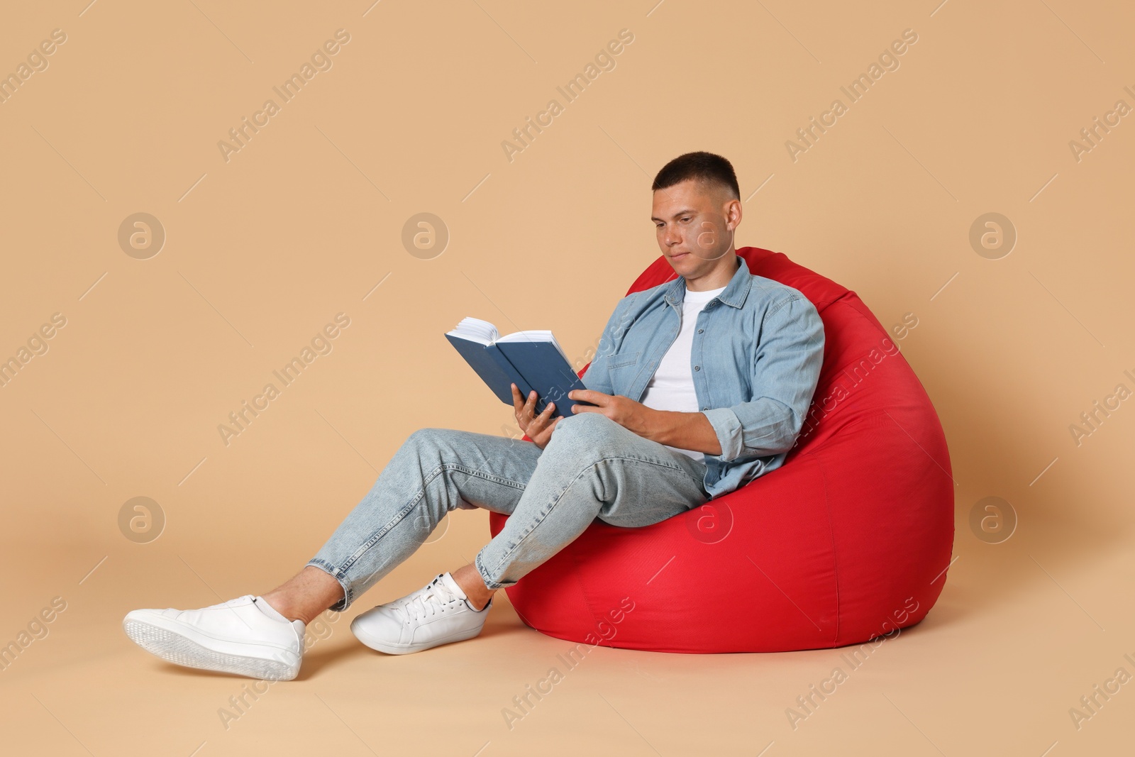 Photo of Handsome man reading book on red bean bag chair against beige background