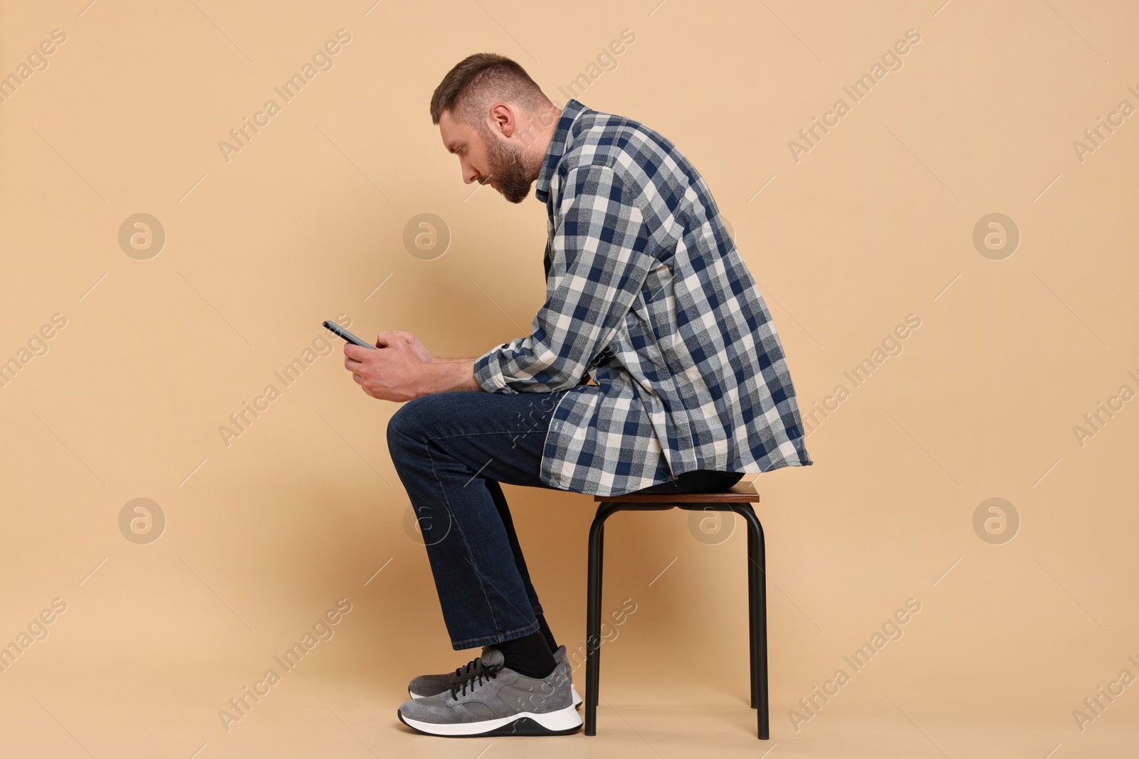 Photo of Man with poor posture sitting on chair and using smartphone against pale orange background