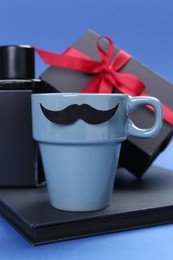 Photo of Happy Father's Day. Cup with paper mustache, notebook, perfume and gift box on blue background, closeup