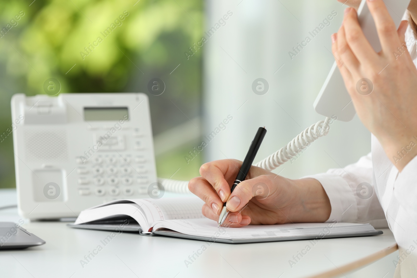 Photo of Assistant with telephone handset writing at white table against blurred green background, closeup