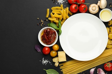 Plate surrounded by different types of pasta, products and spices on black background, flat lay. Space for text