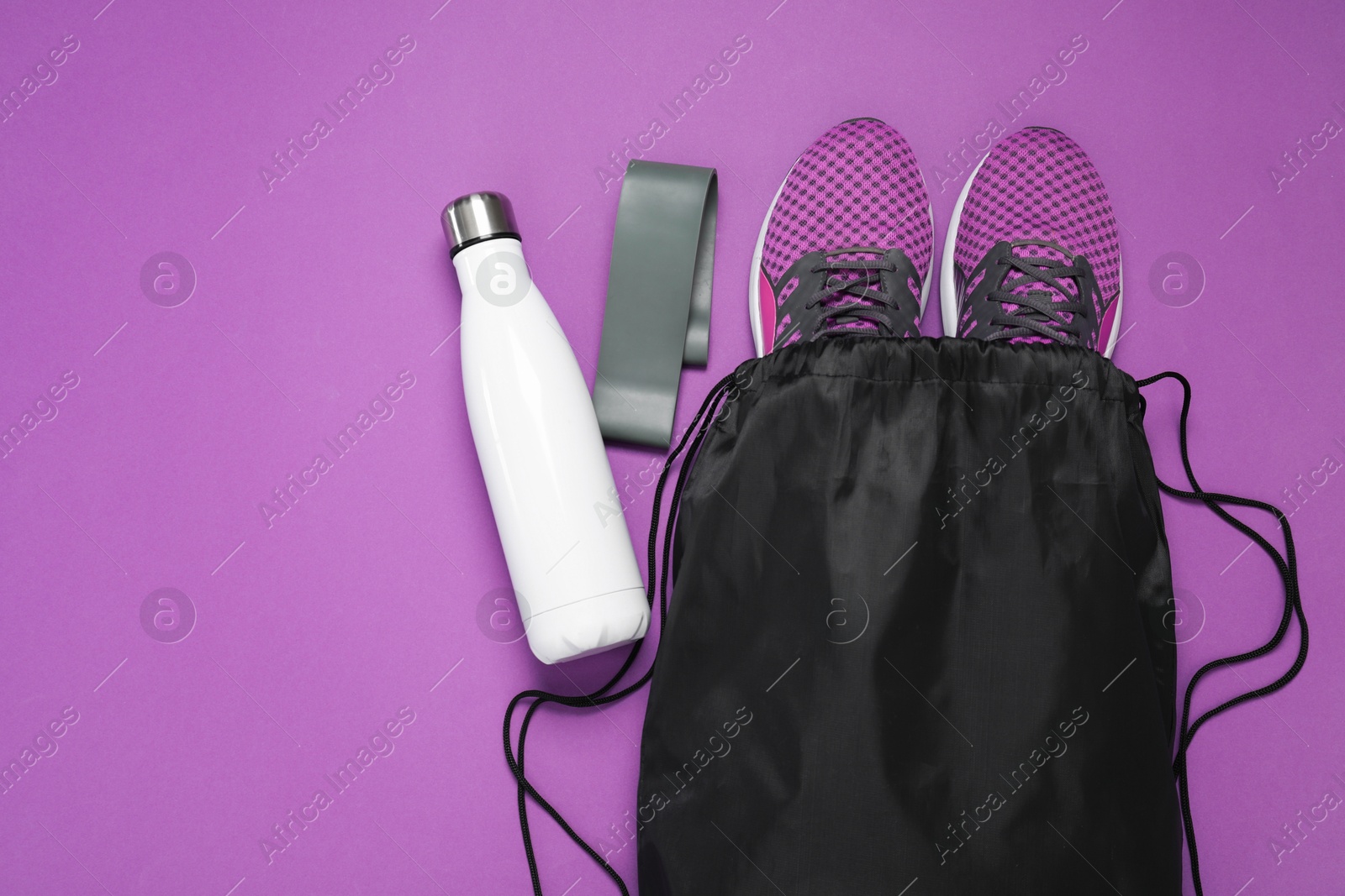 Photo of Black drawstring bag, sneakers, thermo bottle and fitness elastic band on purple background, flat lay. Space for text