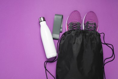 Black drawstring bag, sneakers, thermo bottle and fitness elastic band on purple background, flat lay. Space for text