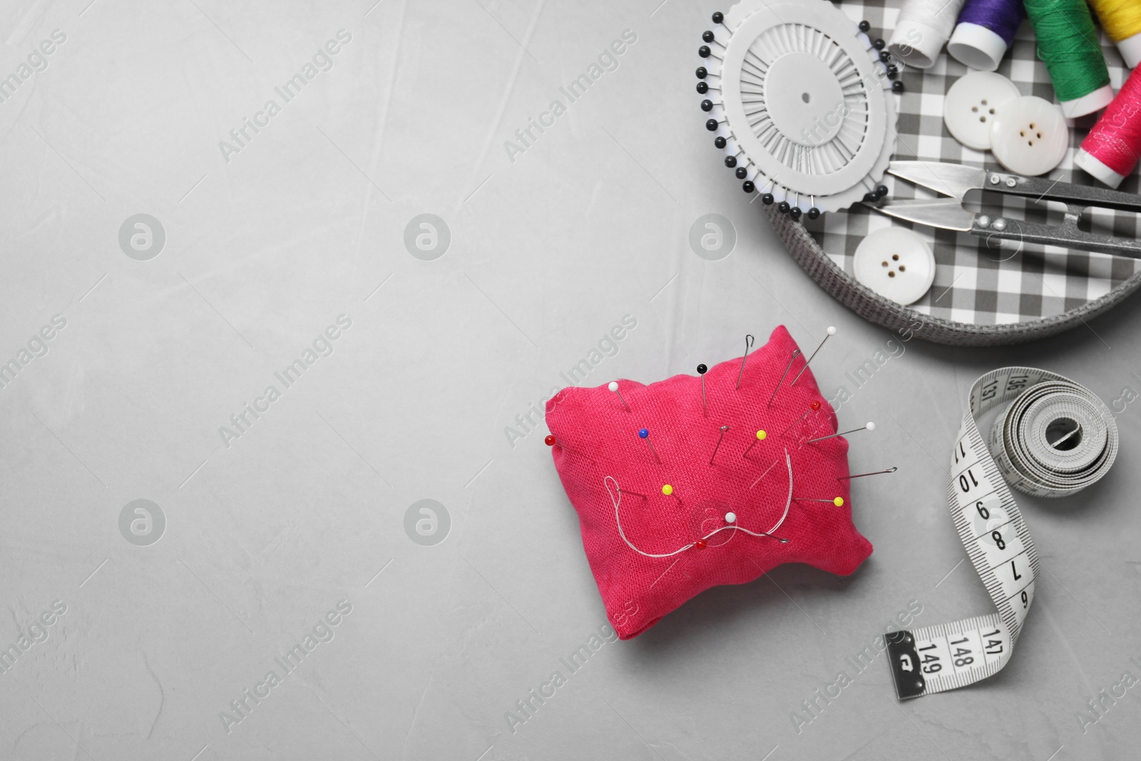 Photo of Red pincushion with pins and other sewing tools on grey table, flat lay. Space for text