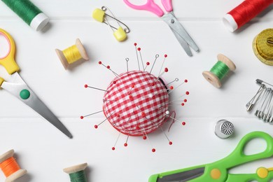 Photo of Checkered pincushion with pins and other sewing tools on white wooden table, flat lay