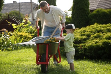 Photo of Father with his son holding wheelbarrow full of beautiful flowers and gardening tools outdoors