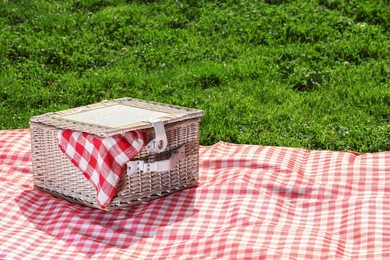 Photo of One picnic wicker basket with checkered napkin and blanket on green grass. Space for text