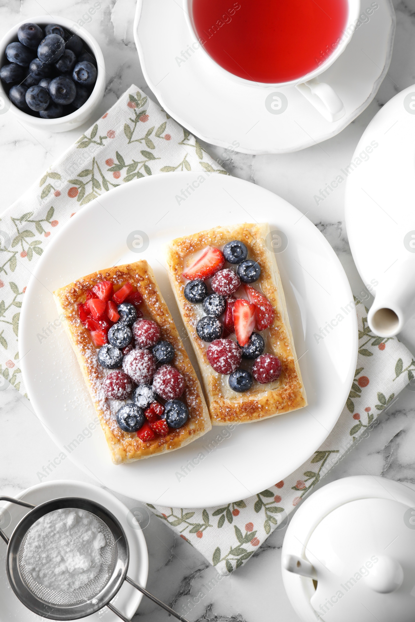 Photo of Tasty puff pastries with berries and tea on white marble table, flat lay