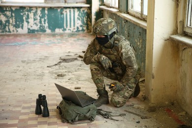 Military mission. Soldier in uniform using laptop and binoculars inside abandoned building