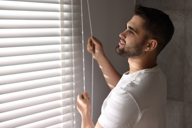 Man adjusting window blinds at home, space for text