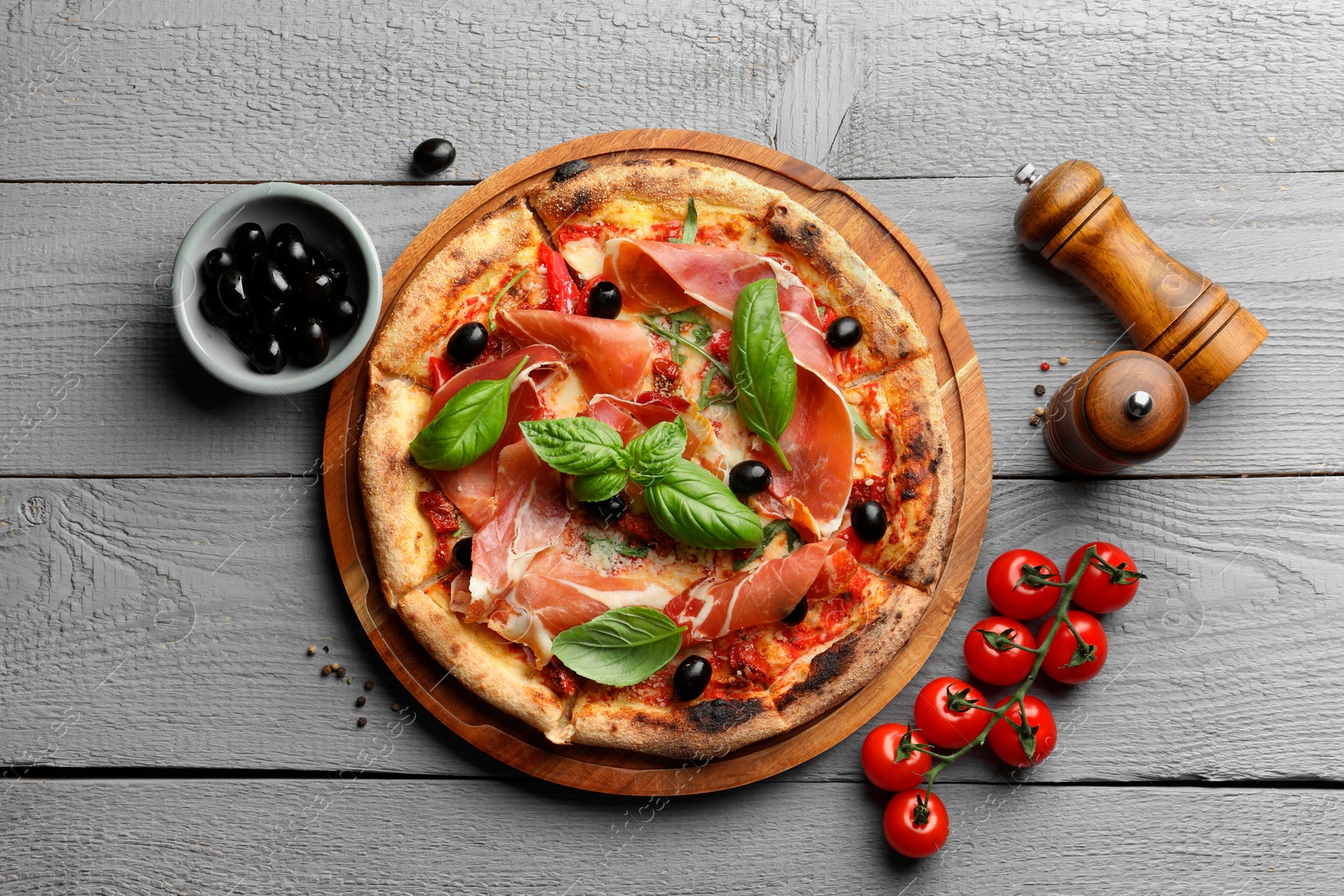 Photo of Tasty pizza with cured ham, olives, tomatoes and basil on gray wooden table, flat lay