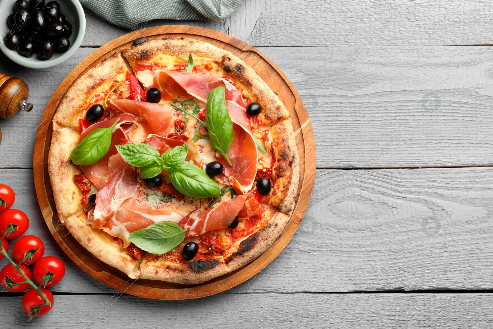 Photo of Tasty pizza with cured ham, olives, tomatoes and basil on gray wooden table, flat lay. Space for text