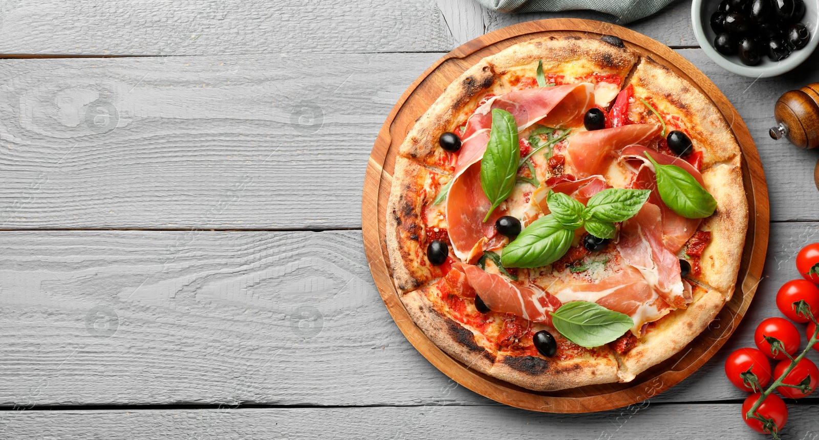 Photo of Tasty pizza with cured ham, olives, tomatoes and basil on gray wooden table, flat lay. Space for text