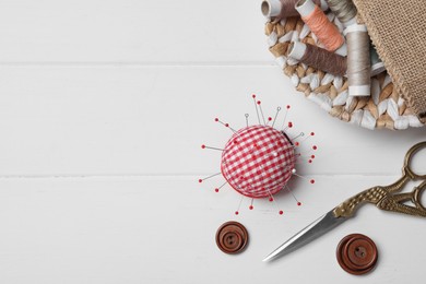 Photo of Checkered pincushion with pins and other sewing tools on white wooden table, flat lay. Space for text