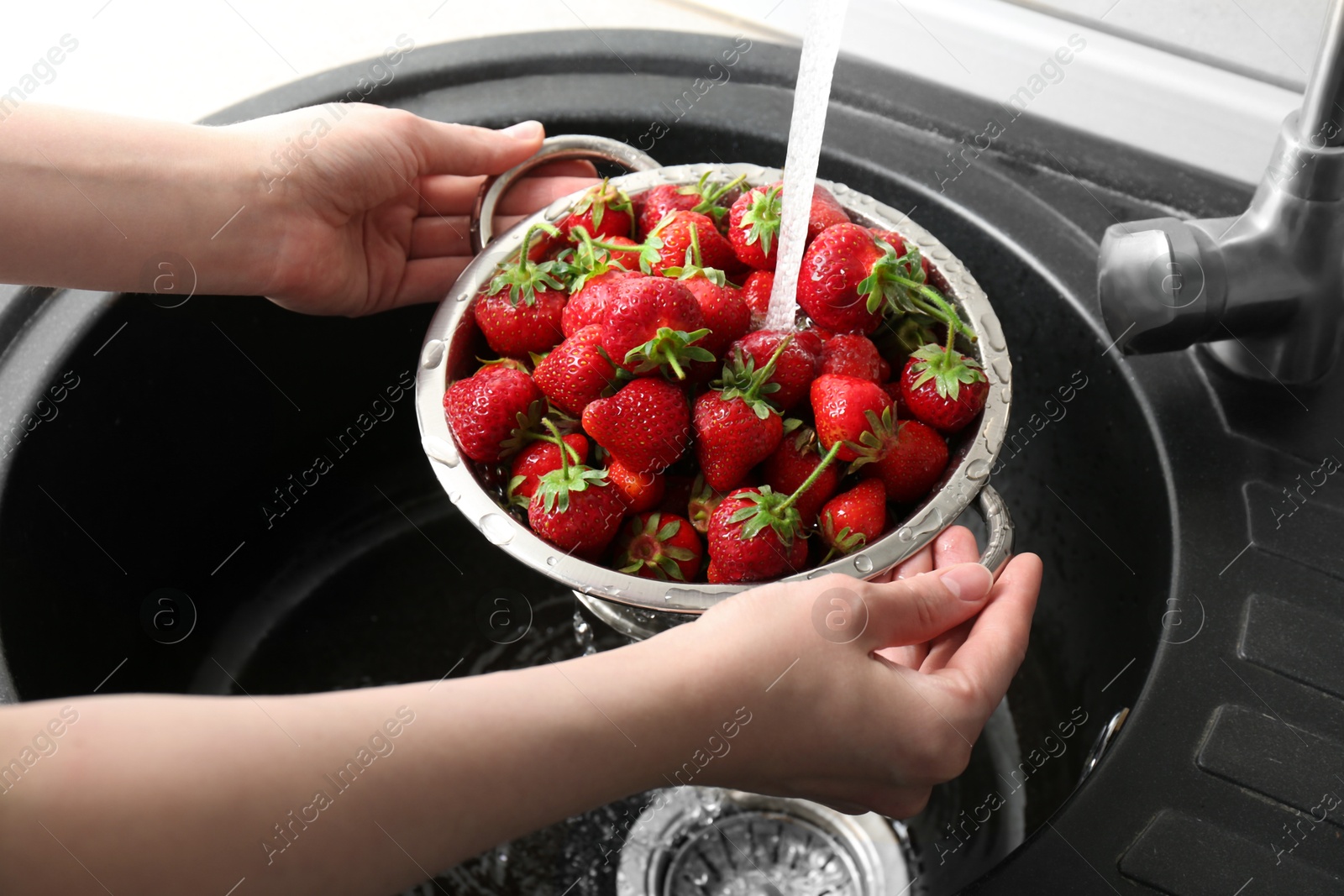 Photo of Woman washing fresh strawberries under tap water in metal colander above sink, closeup