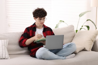 Photo of Teenager taking notes while working with laptop on sofa at home. Remote job