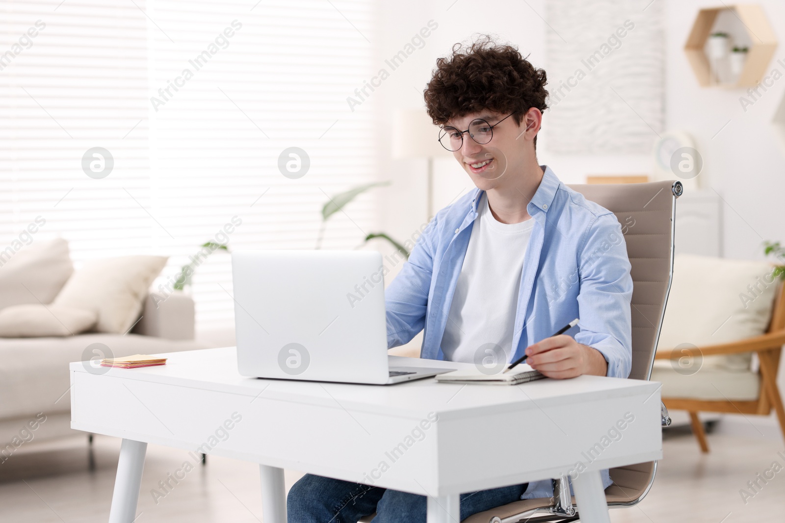 Photo of Teenager taking notes while working with laptop on sofa at home. Remote job