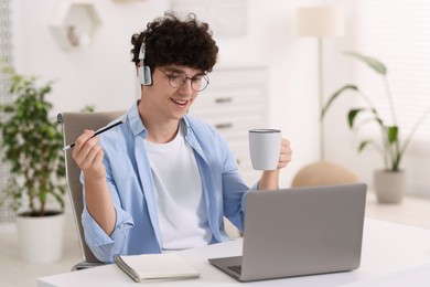 Photo of Teenager with cup of drink and pencil working on laptop at home. Remote job