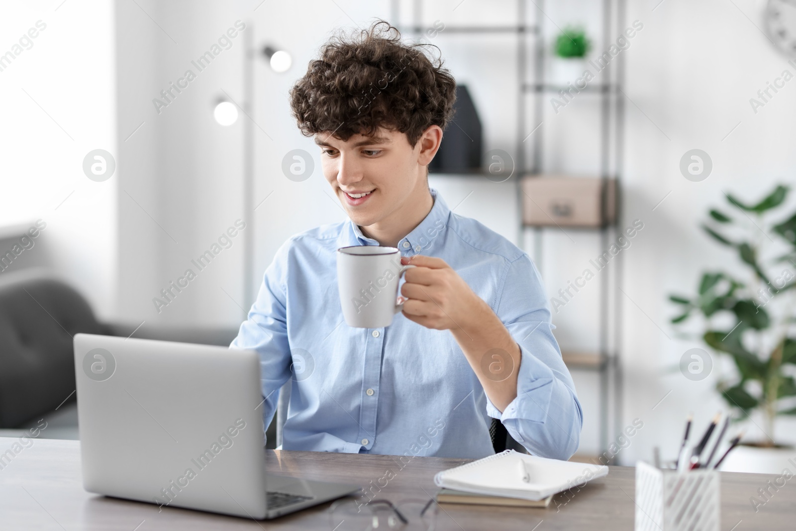 Photo of Teenager with cup of drink working on laptop at table indoors. Remote job