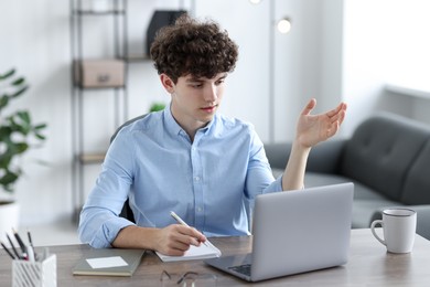 Photo of Teenager taking notes while working with laptop at table indoors. Remote job