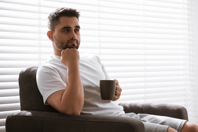Photo of Man with cup of drink sitting on armchair near window blinds indoors