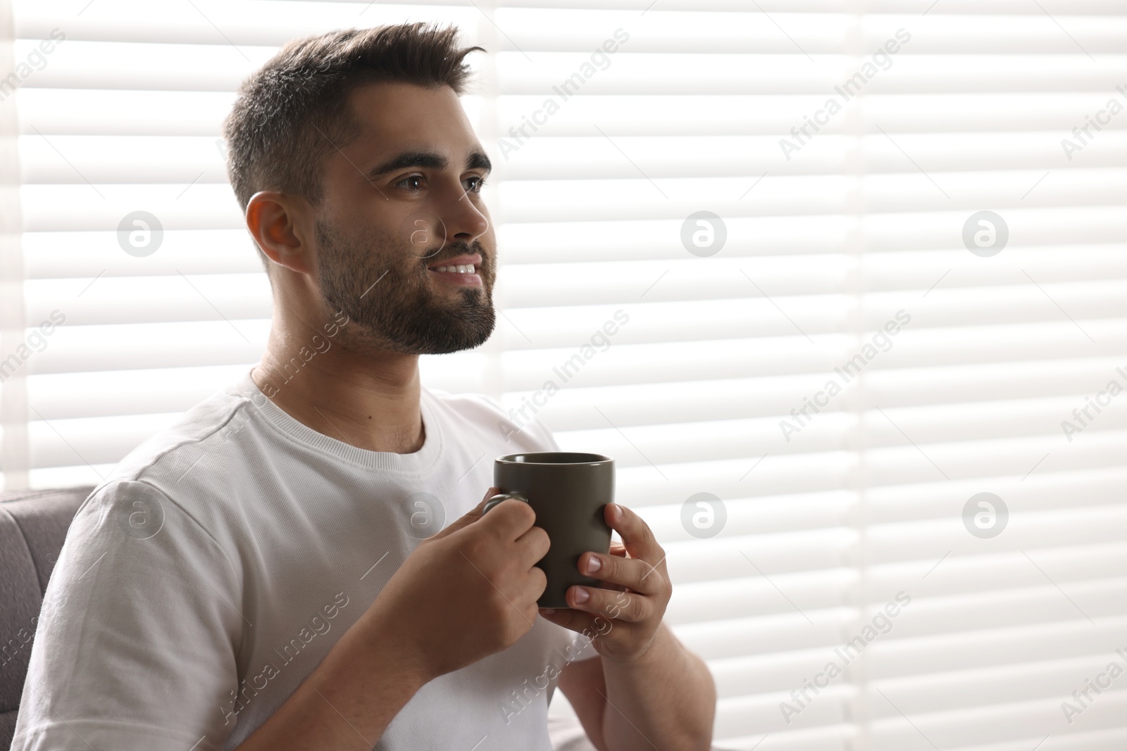 Photo of Man with cup of drink sitting on armchair near window blinds indoors, space for text