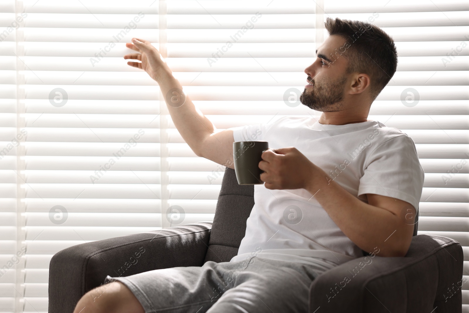 Photo of Man with cup of drink sitting on armchair near window blinds indoors