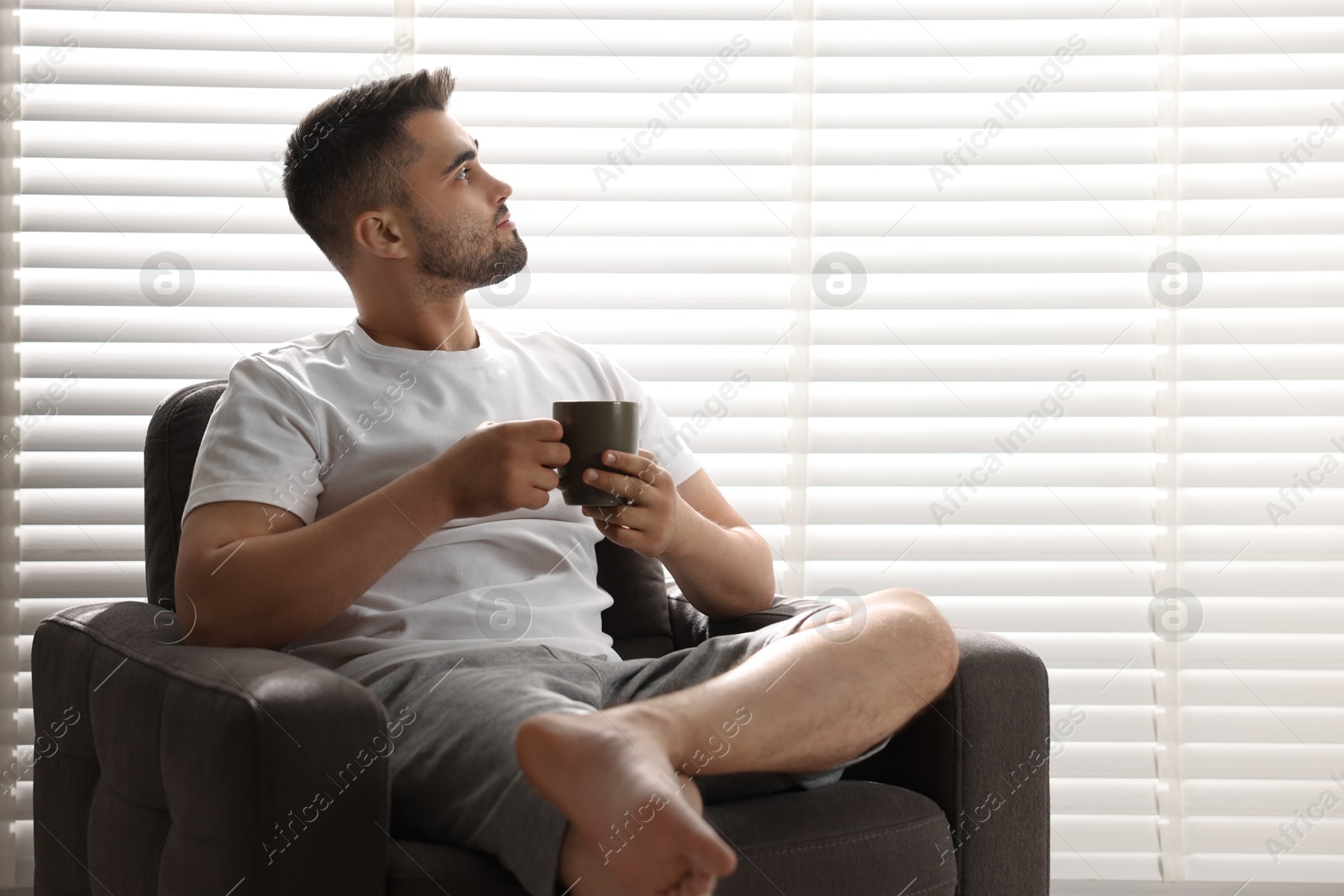 Photo of Man with cup of drink sitting on armchair near window blinds indoors