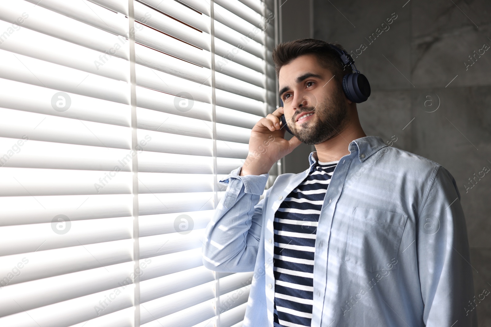 Photo of Man with headphones listening to music near window blinds at home, space for text