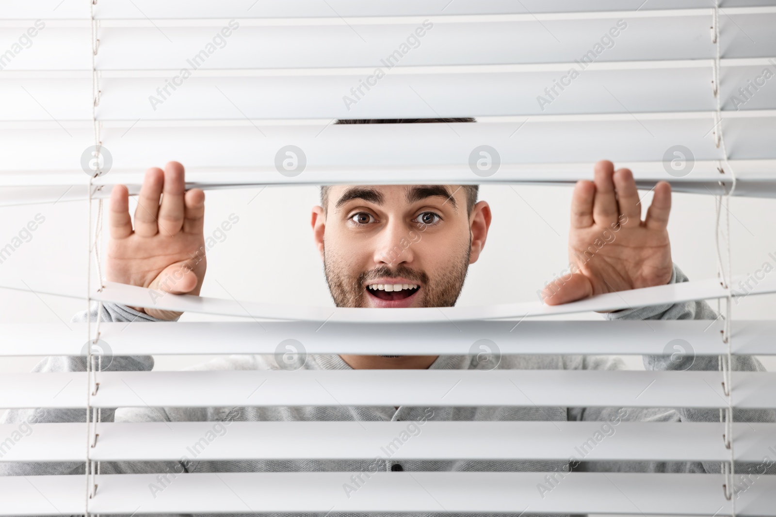 Photo of Young man looking through window blinds on white background