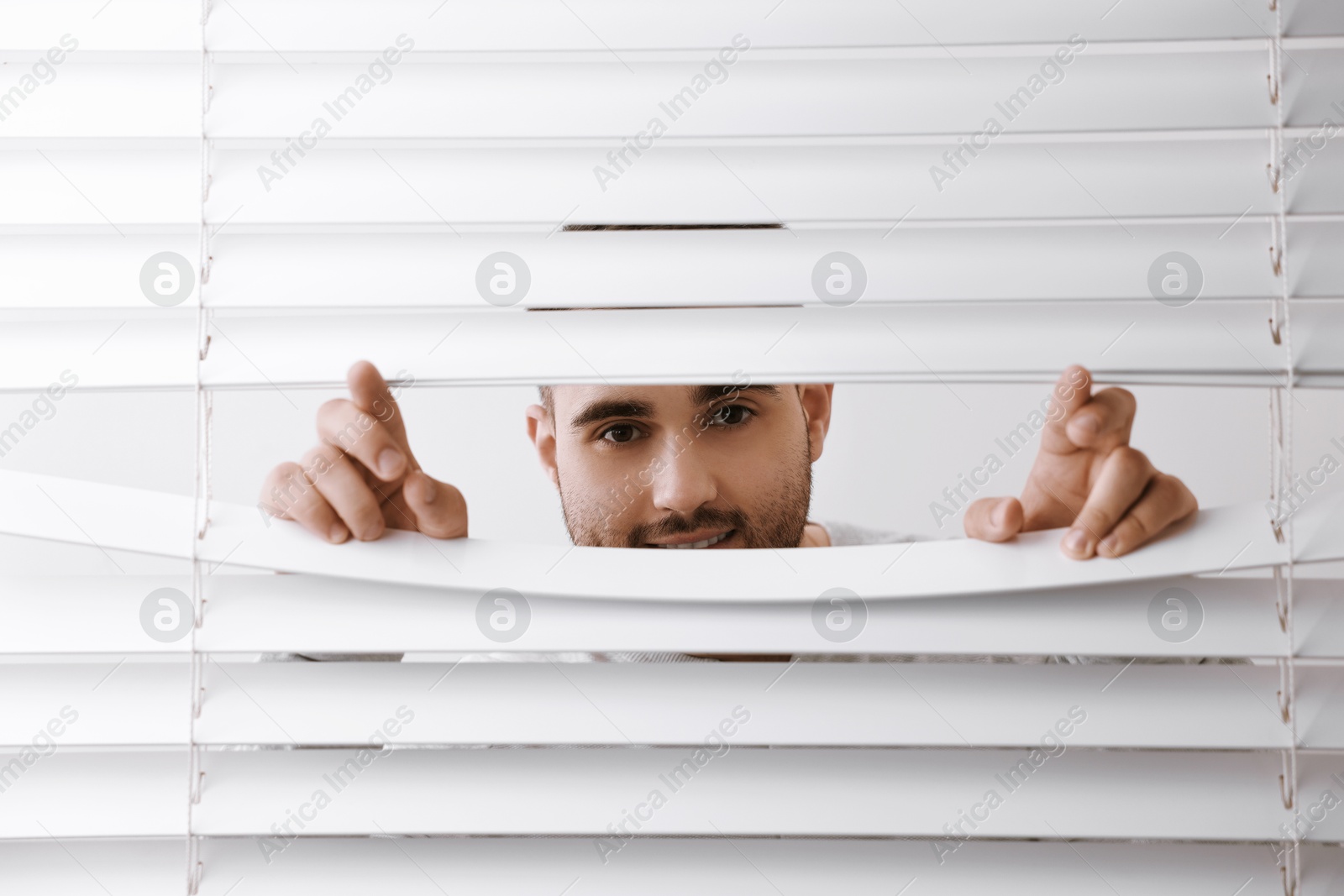 Photo of Young man looking through window blinds on white background