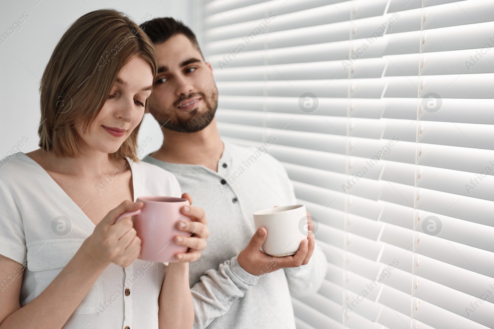 Photo of Couple with cups of drink near window blinds at home, space for text