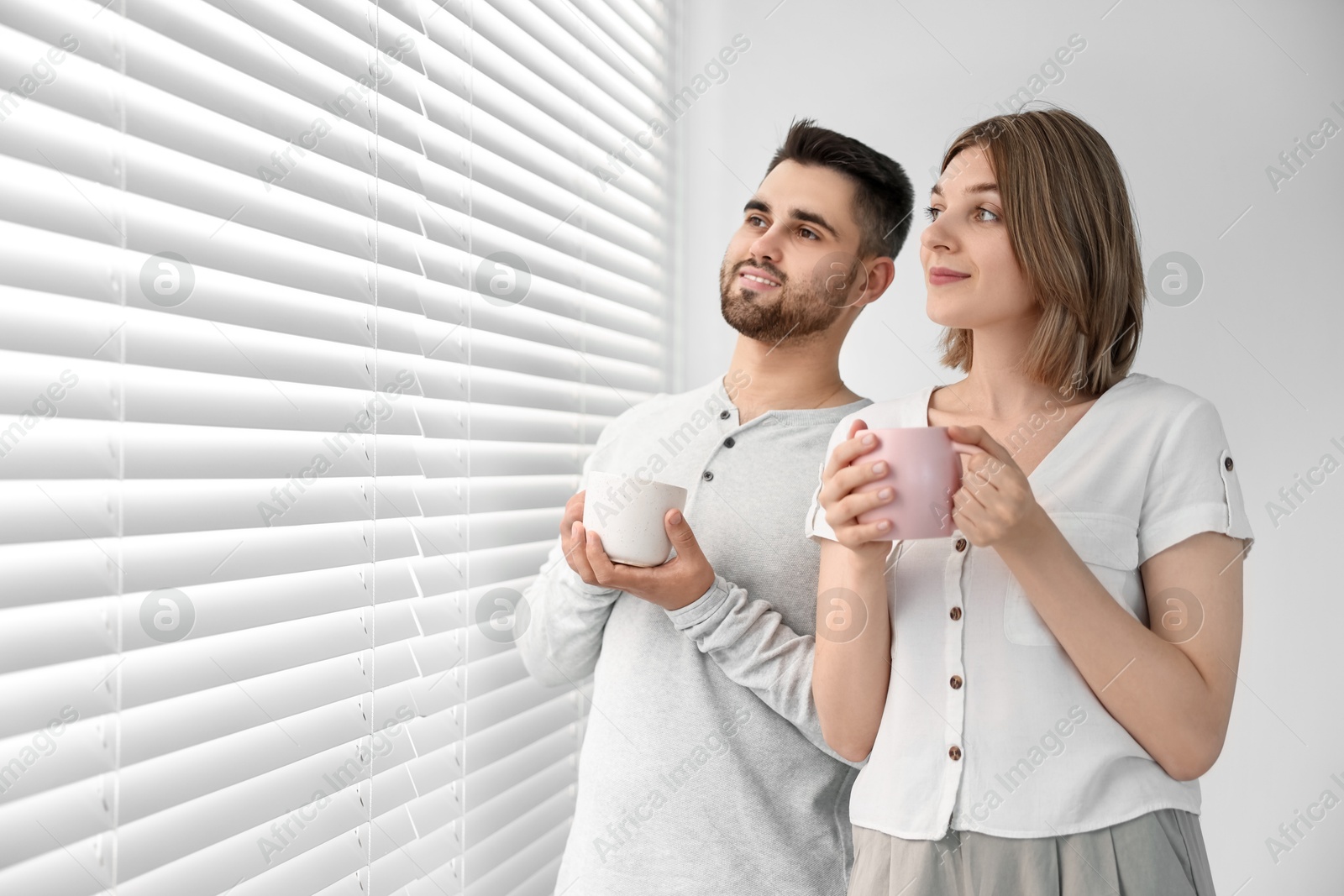 Photo of Couple with cups of drink near window blinds at home, space for text