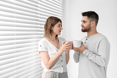 Photo of Couple with cups of drink near window blinds at home