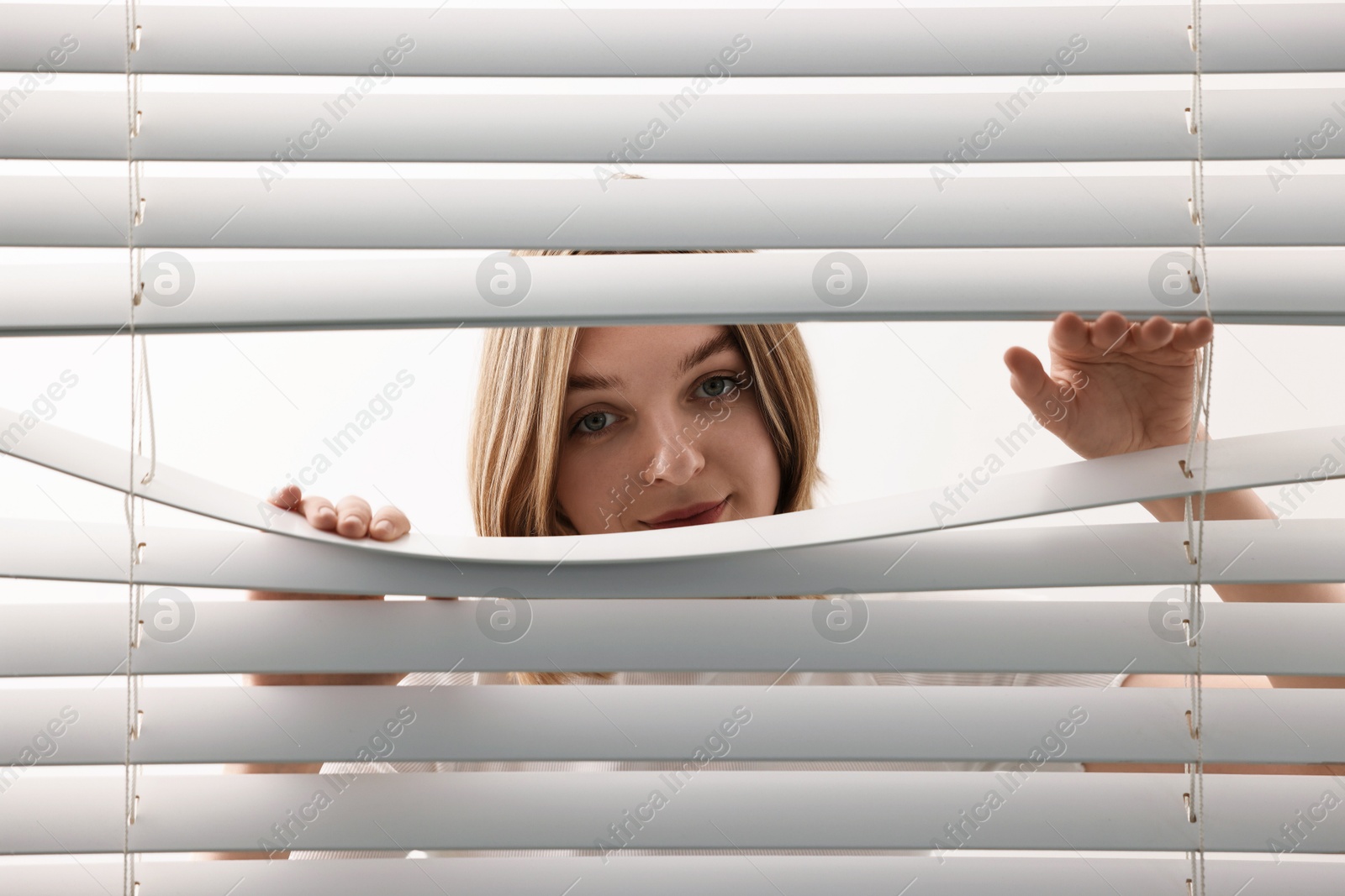 Photo of Young woman looking through window blinds on white background