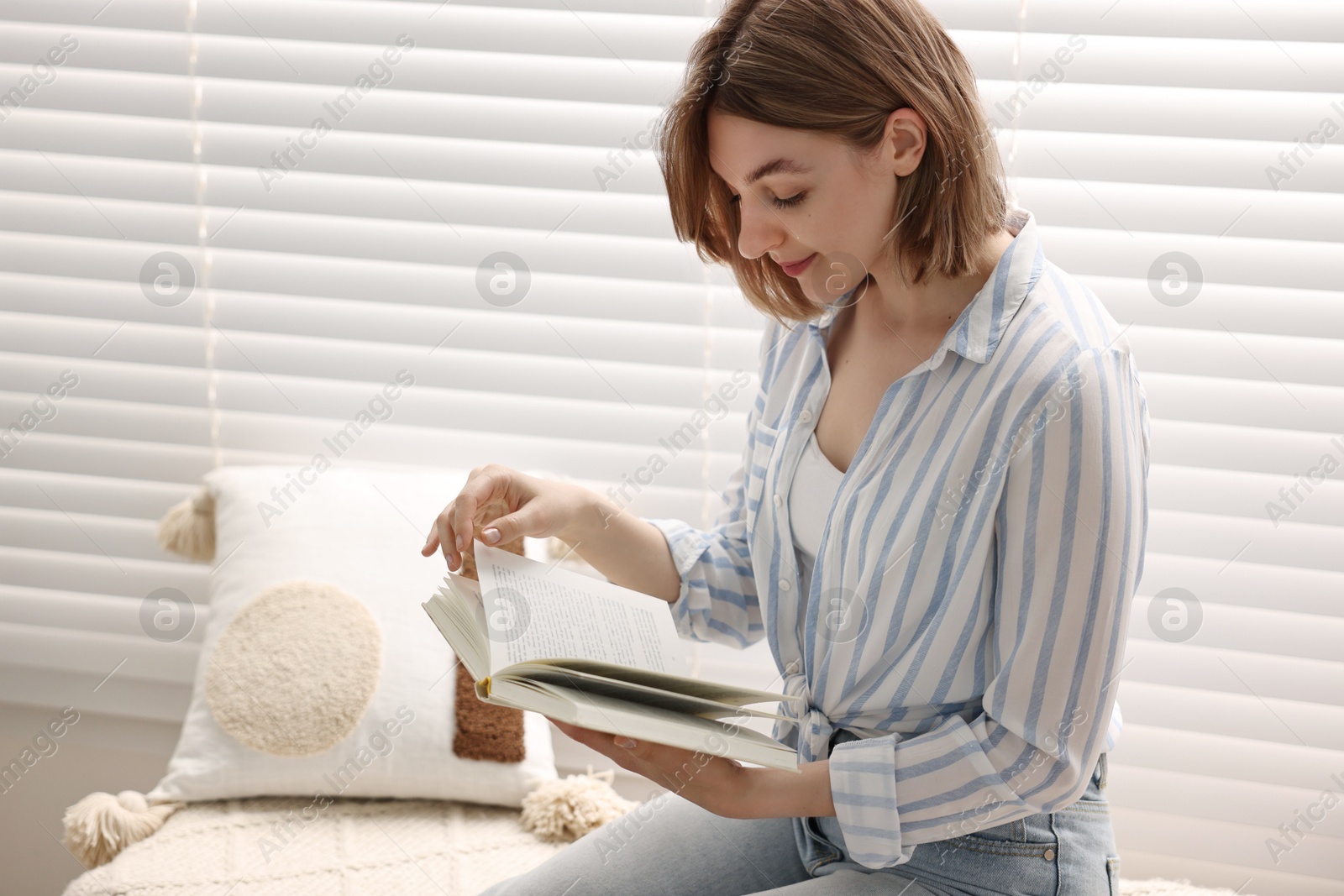 Photo of Woman reading book near window blinds at home