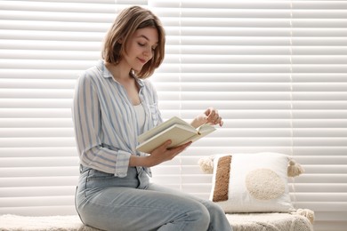 Photo of Woman reading book near window blinds at home