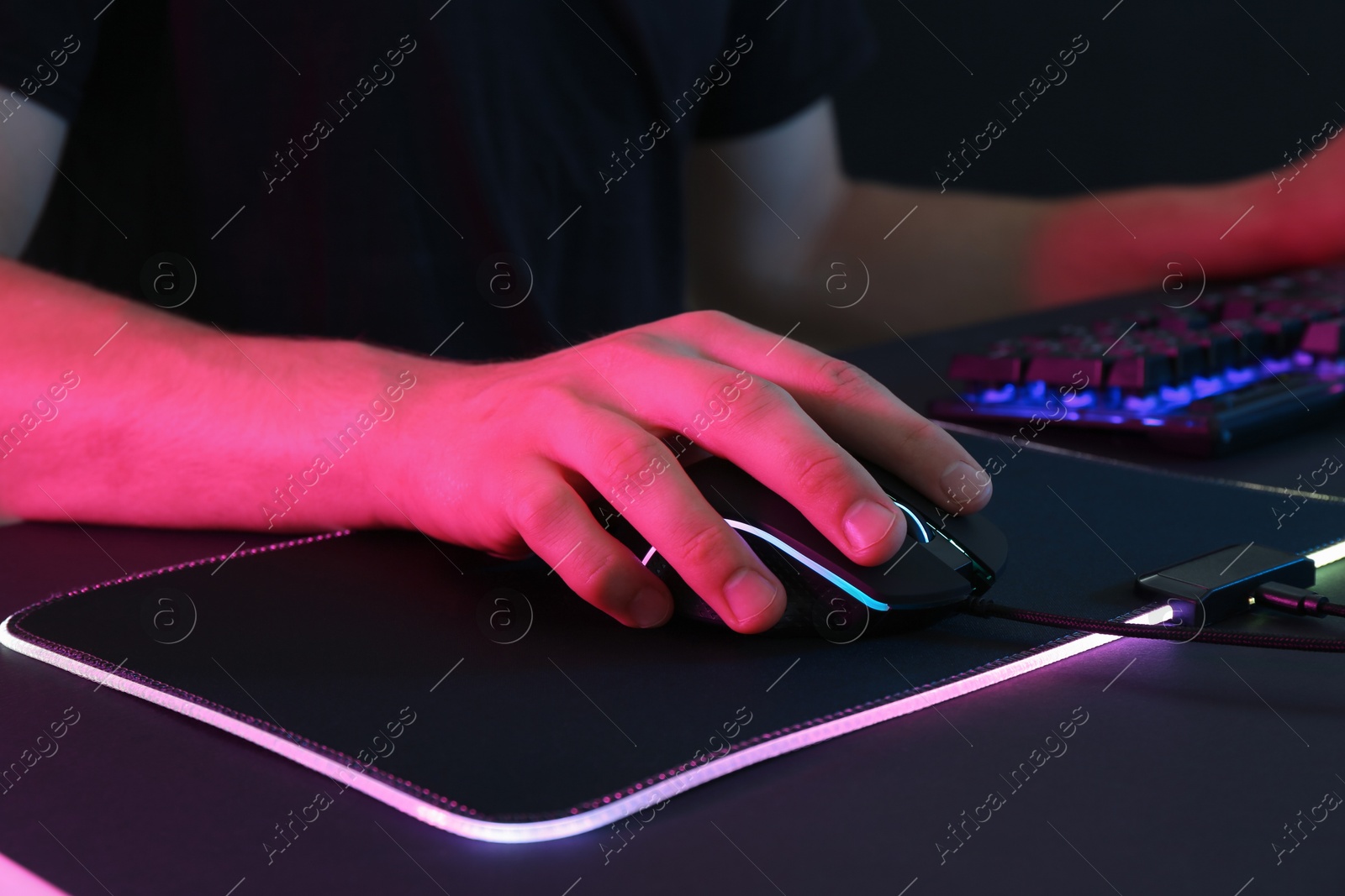 Photo of Gamer using modern wired computer mouse in neon lights at black table, closeup