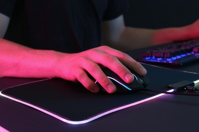 Gamer using modern wired computer mouse in neon lights at black table, closeup