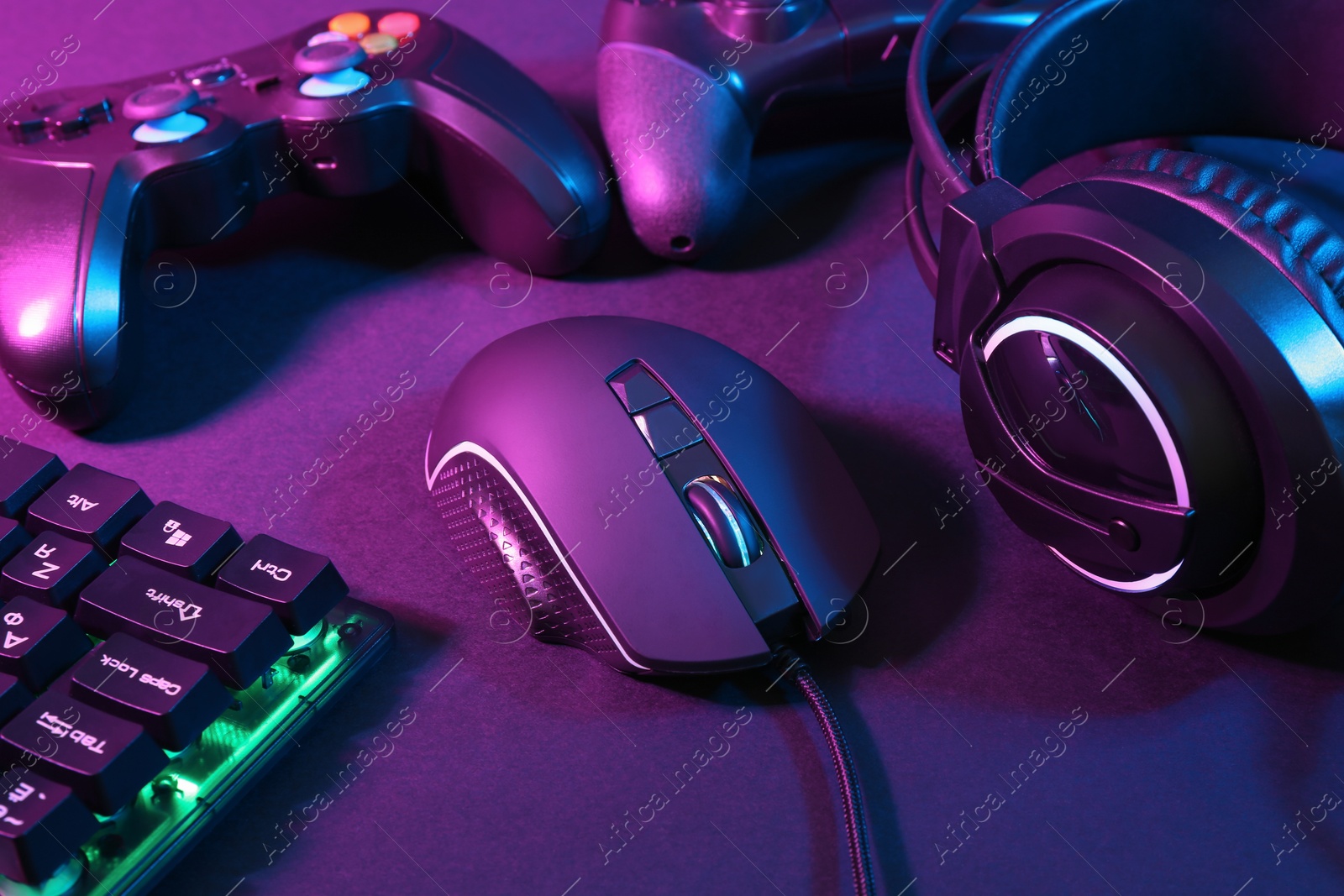 Photo of Computer mouse, headset, RGB keyboard and game controllers in neon lights on dark background, closeup