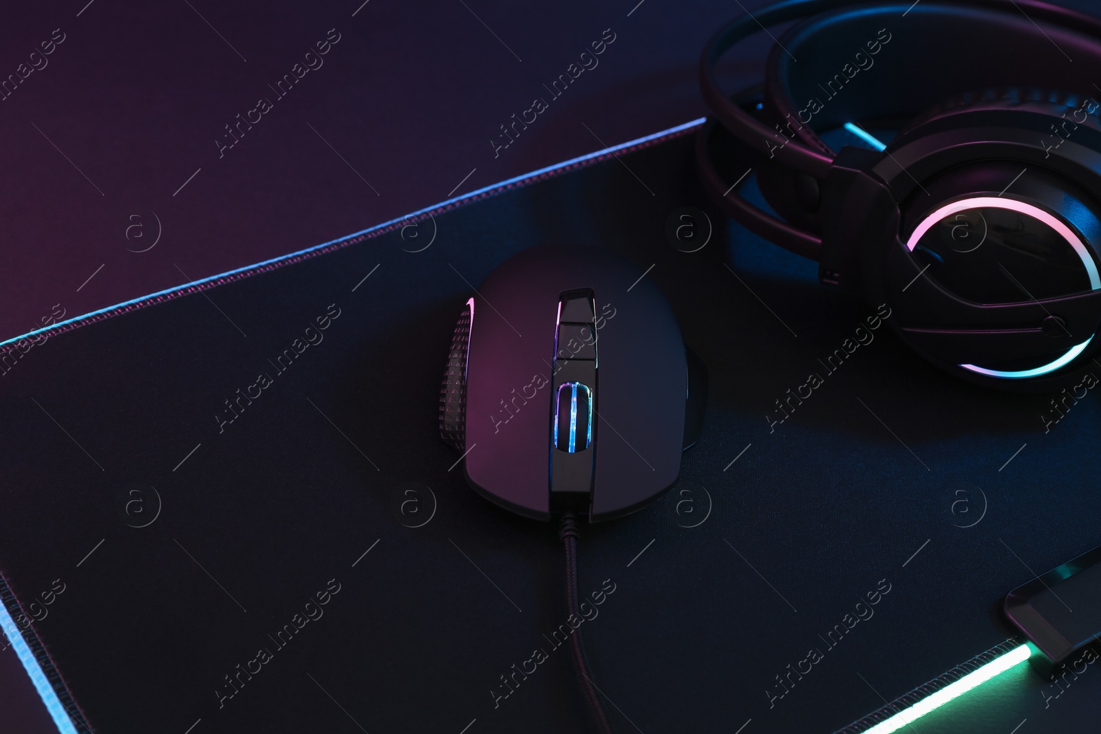 Photo of Computer mouse, mousepad and headset in neon lights on dark background, closeup