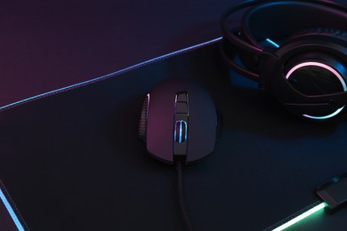 Photo of Computer mouse, mousepad and headset in neon lights on dark background, closeup