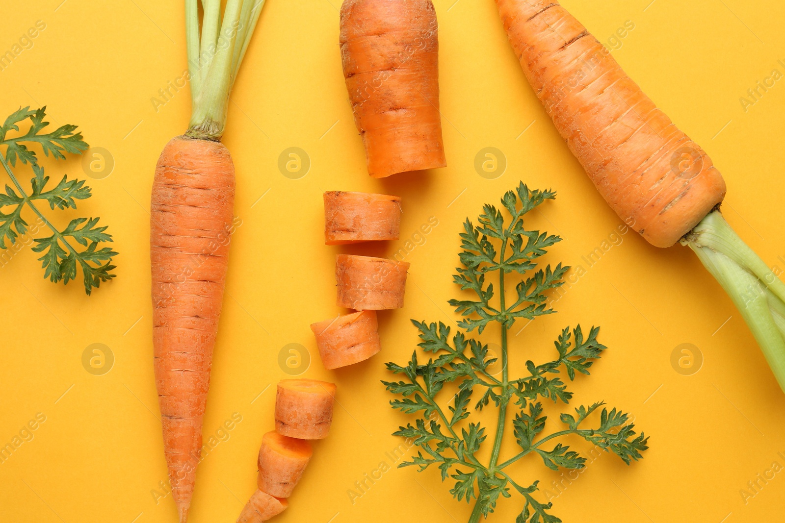 Photo of Whole and cut fresh carrots with green leaves on yellow background, flat lay