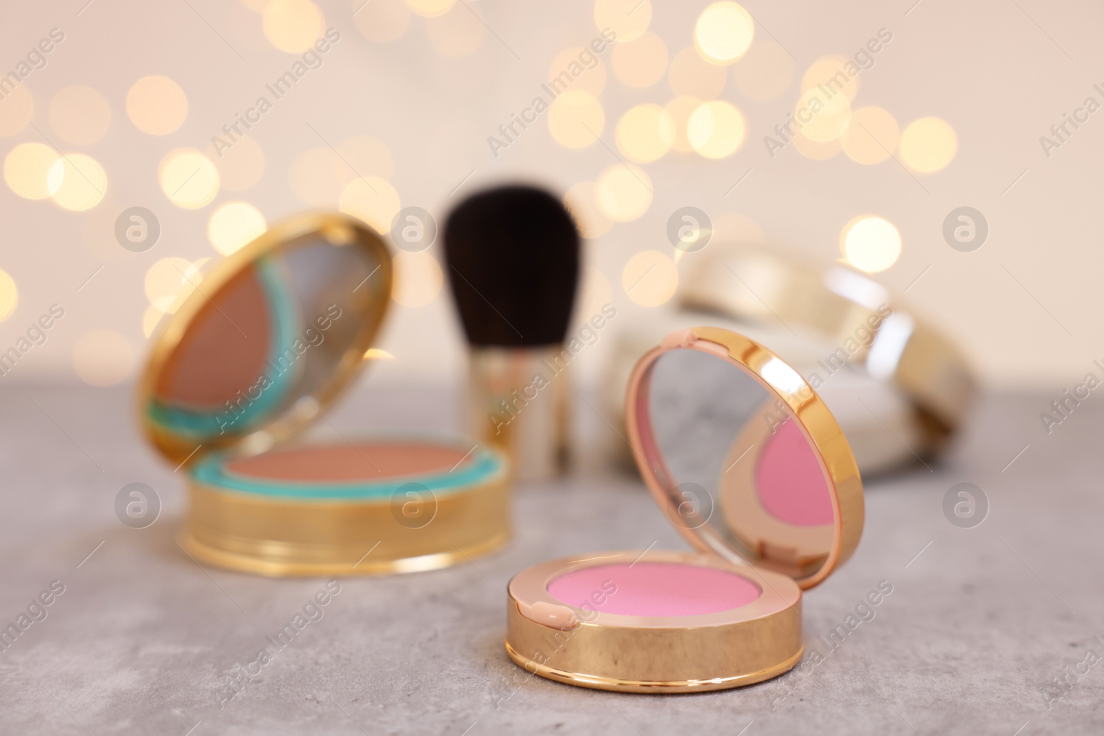 Photo of Face blusher and other cosmetic products on grey textured table against blurred lights, closeup