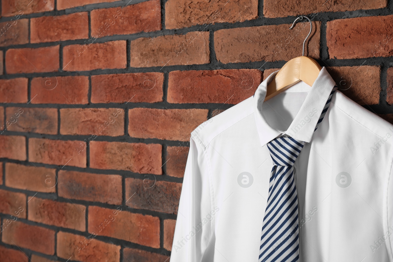 Photo of Hanger with white shirt and striped necktie on red brick wall, space for text