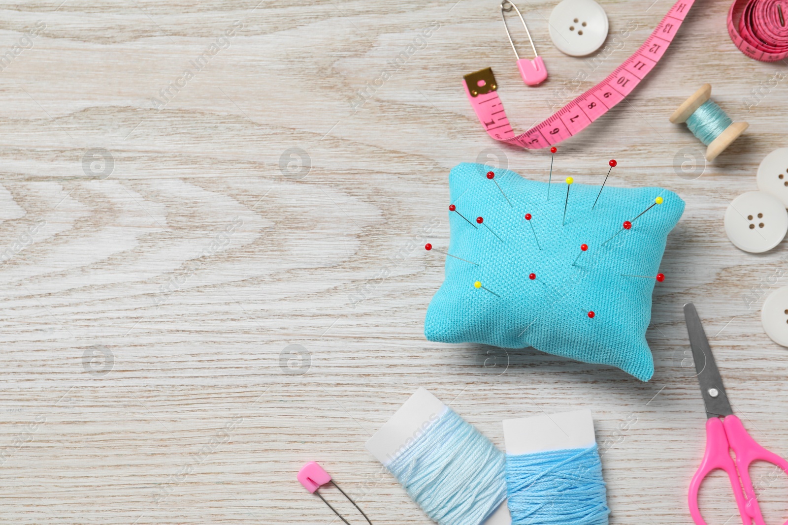 Photo of Light blue pincushion with pins and other sewing tools on wooden table, flat lay. Space for text