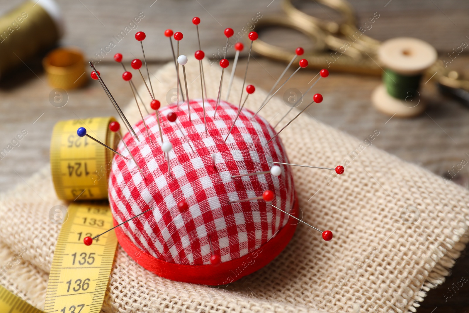 Photo of Checkered pincushion with pins and other sewing tools on wooden table, closeup