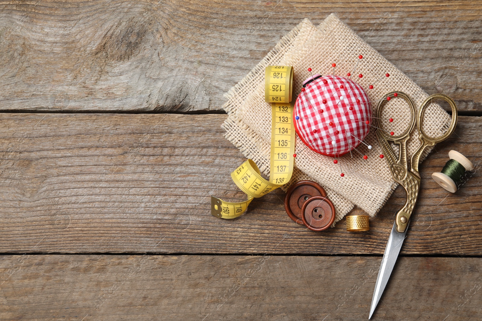 Photo of Checkered pincushion with pins and other sewing tools on wooden table, flat lay. Space for text