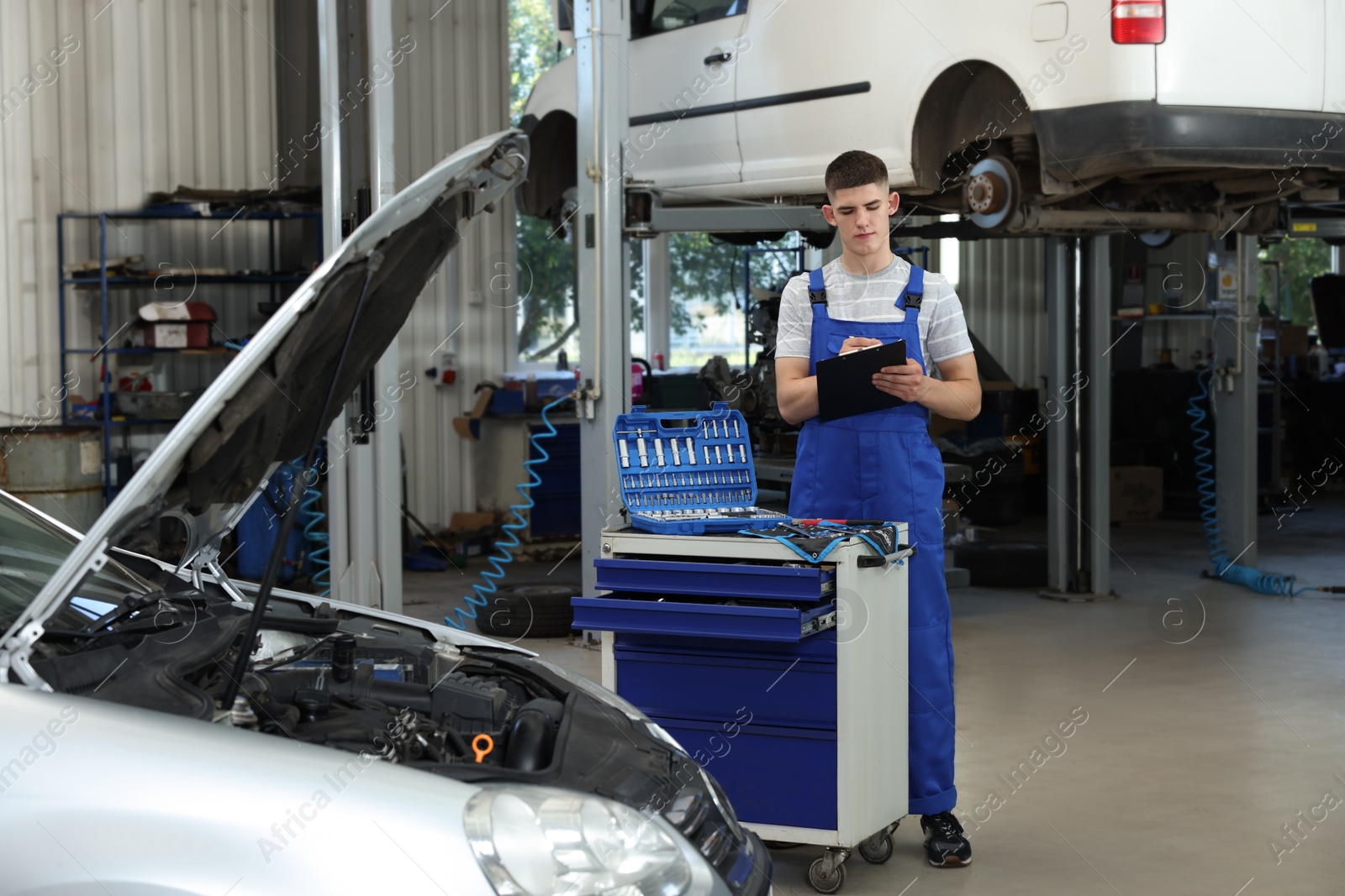 Photo of Young auto mechanic with clipboard taking notes near broken car at automobile repair shop