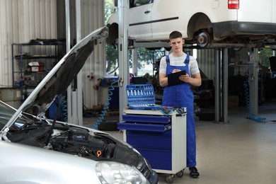 Photo of Young auto mechanic with clipboard taking notes near broken car at automobile repair shop